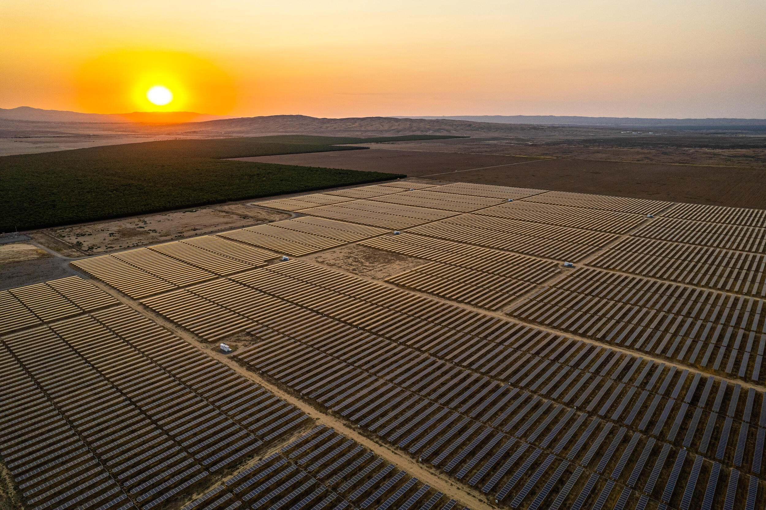 Aerial view of a farm of solar panels in a semi-arid setting with sun setting over a ridge in the distance.